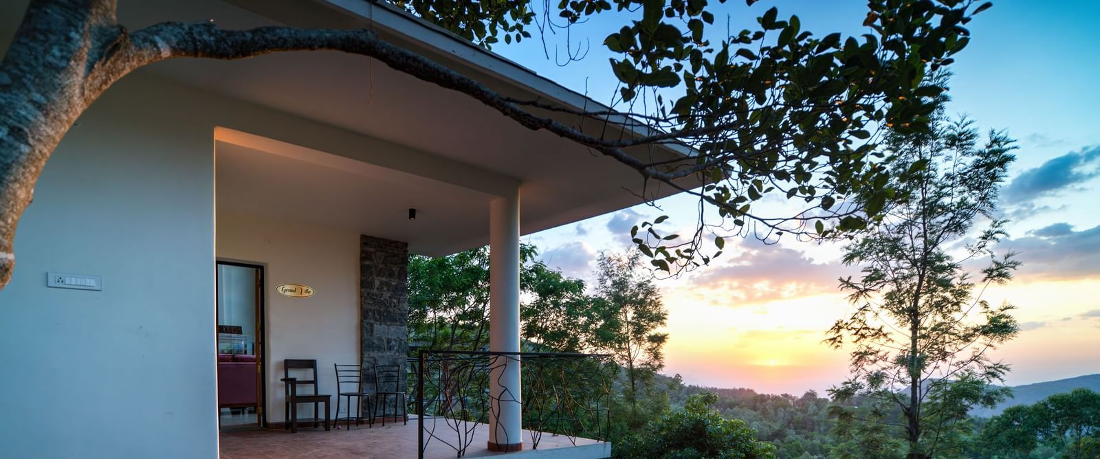 A curved balcony of a hotel room with a tiled floor and a small seating area, overlooking a lush green forest at sunset