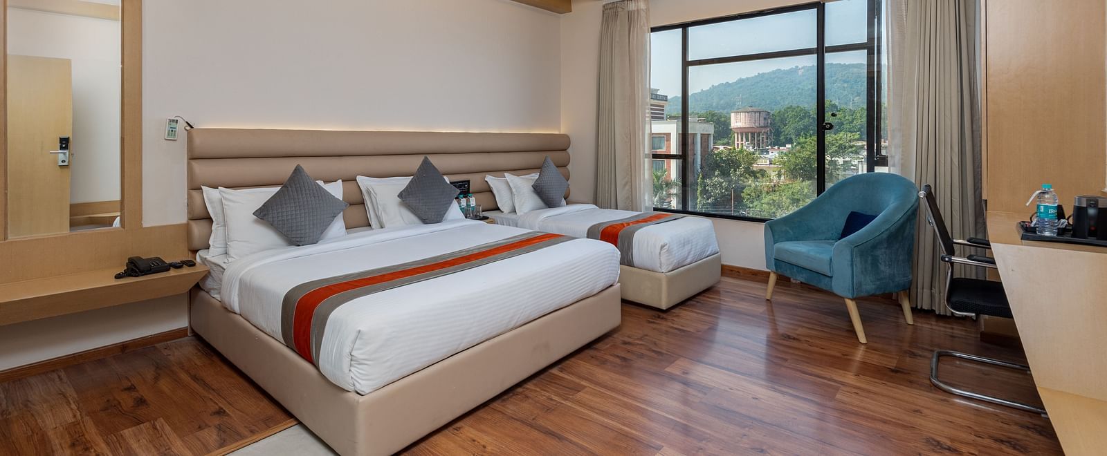 An image of a room with a king size bed and a single bed along with a chair, and a glass window overlooking the city view - DLS Hotel Arihant, Haridwar