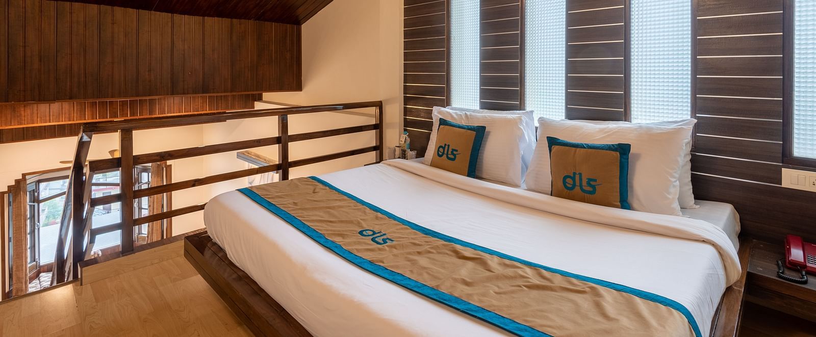 A bedroom with a wooden ceiling, a king size bed with neatly arranged pillows, and a modern design. The room features large windows with blinds and soft lighting - DLS Meghavan Resort, Dharamshala