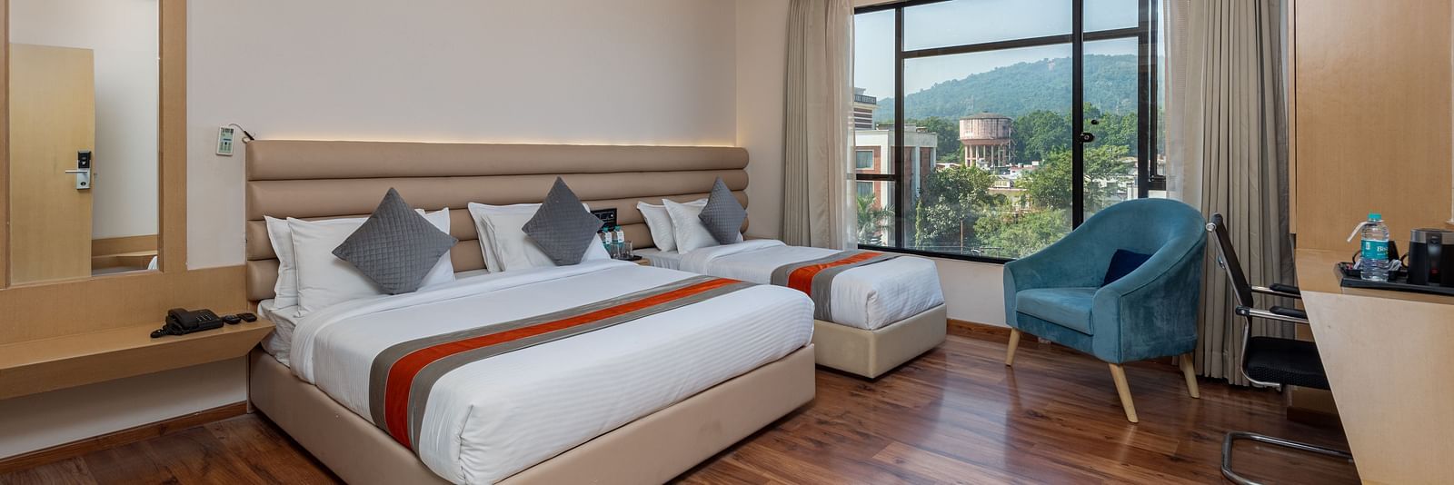 An image of a room with a king size bed and a single bed along with a chair, and a glass window overlooking the city view - DLS Hotel Arihant, Haridwar