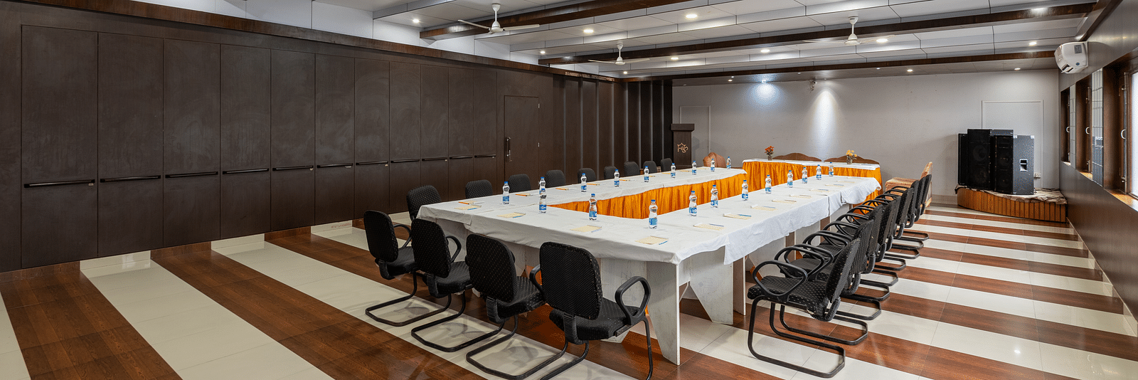 A conference room with a long table, chairs, and decorative flowers. The room features wooden panelling, bright lighting, ceiling fans, and windows along the walls - DLS Meghavan Resort, Dharamshala