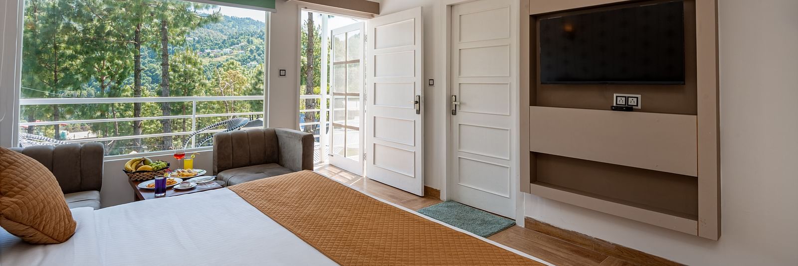 A bright hotel room with a double bed, TV, and seating area, opening onto a balcony with scenic hill views - DLS Nature Trinket Resort, Dalhousie.