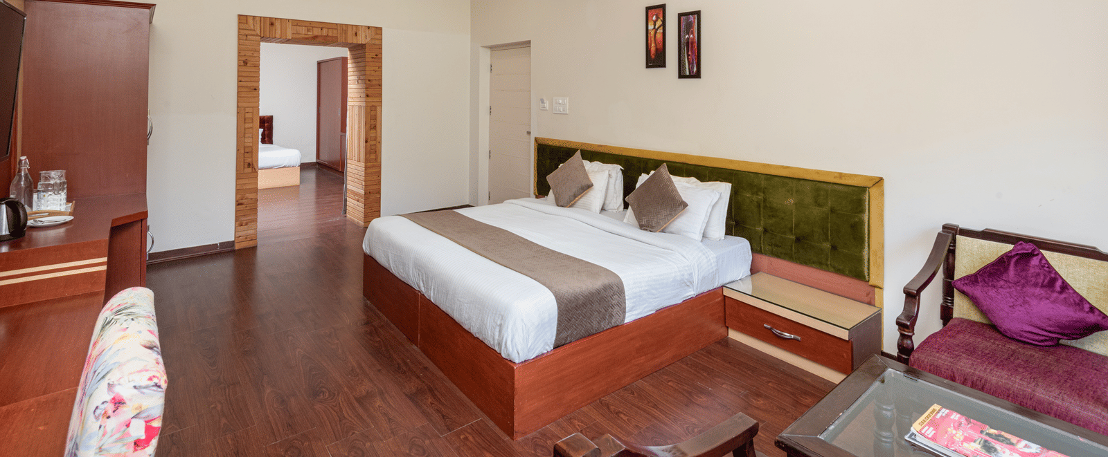 The image shows a spacious hotel room with a large bed, seating area, and desk. It's decorated with artwork and has a ceiling fan | DLS Tehri Club Resort