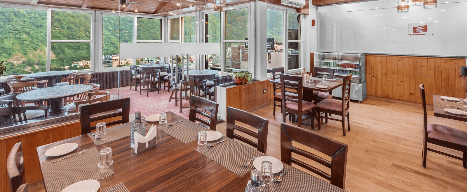 A dining area at DLS Hotels on the Ganges, Rishikesh with wooden tables and chairs, ceiling lights, and large windows showing greenery outside.