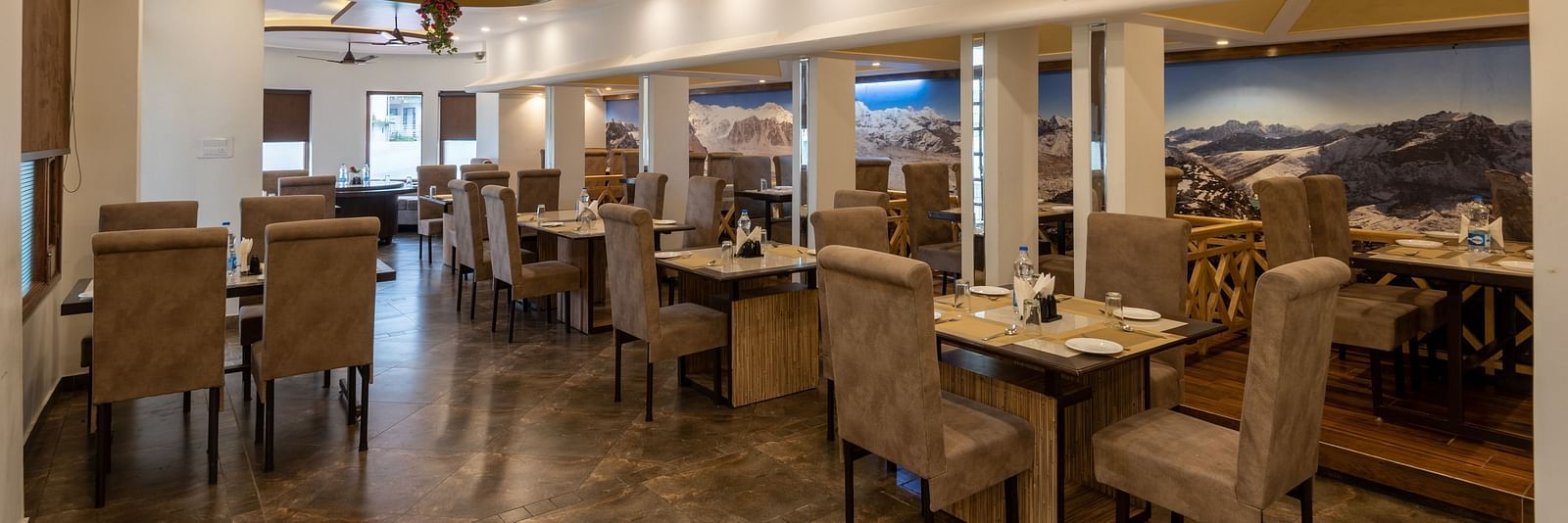 A spacious restaurant in Bhagsu with a modern design, featuring large mountain murals on the walls and rows of tables and chairs – DLS Meghavan Resort.