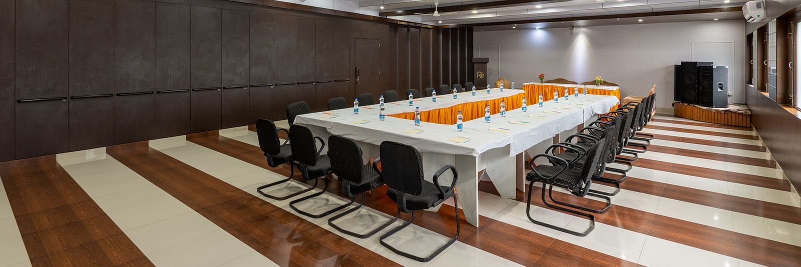 A conference room with a long table, chairs, and decorative flowers. The room features wooden panelling, bright lighting, ceiling fans, and windows along the walls - DLS Meghavan Resort, Dharamshala
