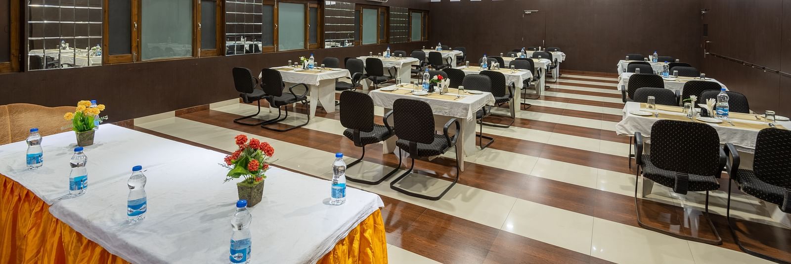 A conference hall in Bhagsu with arranged tables, chairs, and decorative flowers. The room features wooden panelling, bright lights, and large windows - DLS Meghavan Resort, Dharamshala