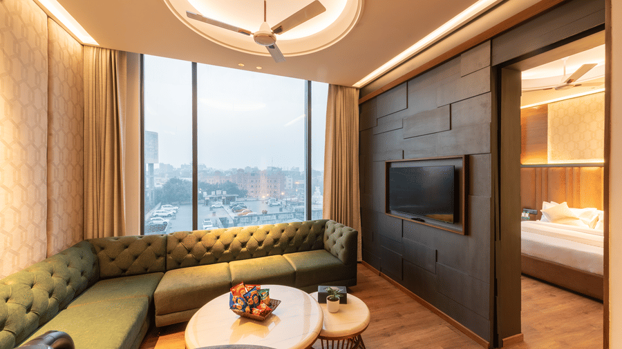 A living room with a sofa, wall-mounted TV unit, table, and a bed visible in the background at DLS Hotels Amritsar.