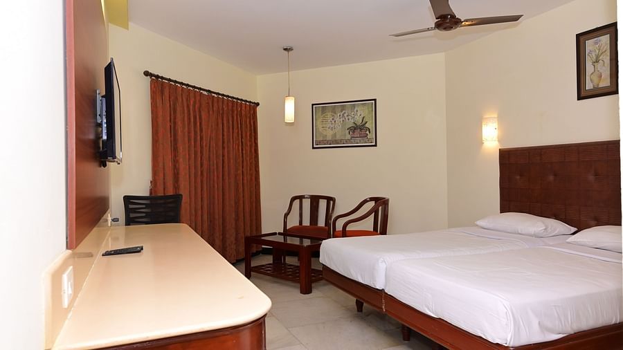 Standard Room with plush bed in front of a TV mounted on the wall and a sitting space by the window featuring two wooden chairs at Jayaram Hotel, Pondicherry