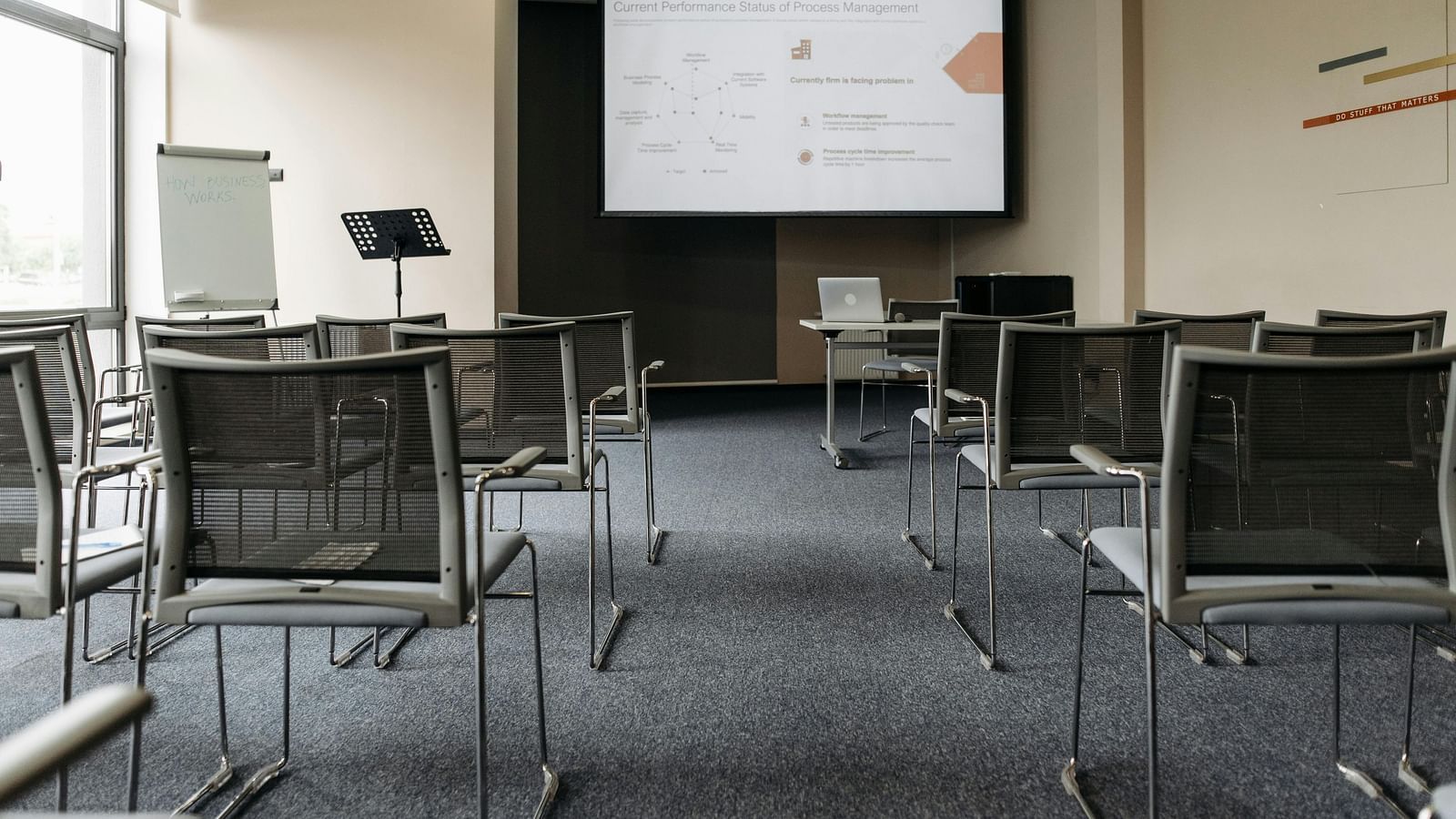 Empty conference room with grey mesh chairs facing a projection screen, a flip chart, and natural light from a large window.