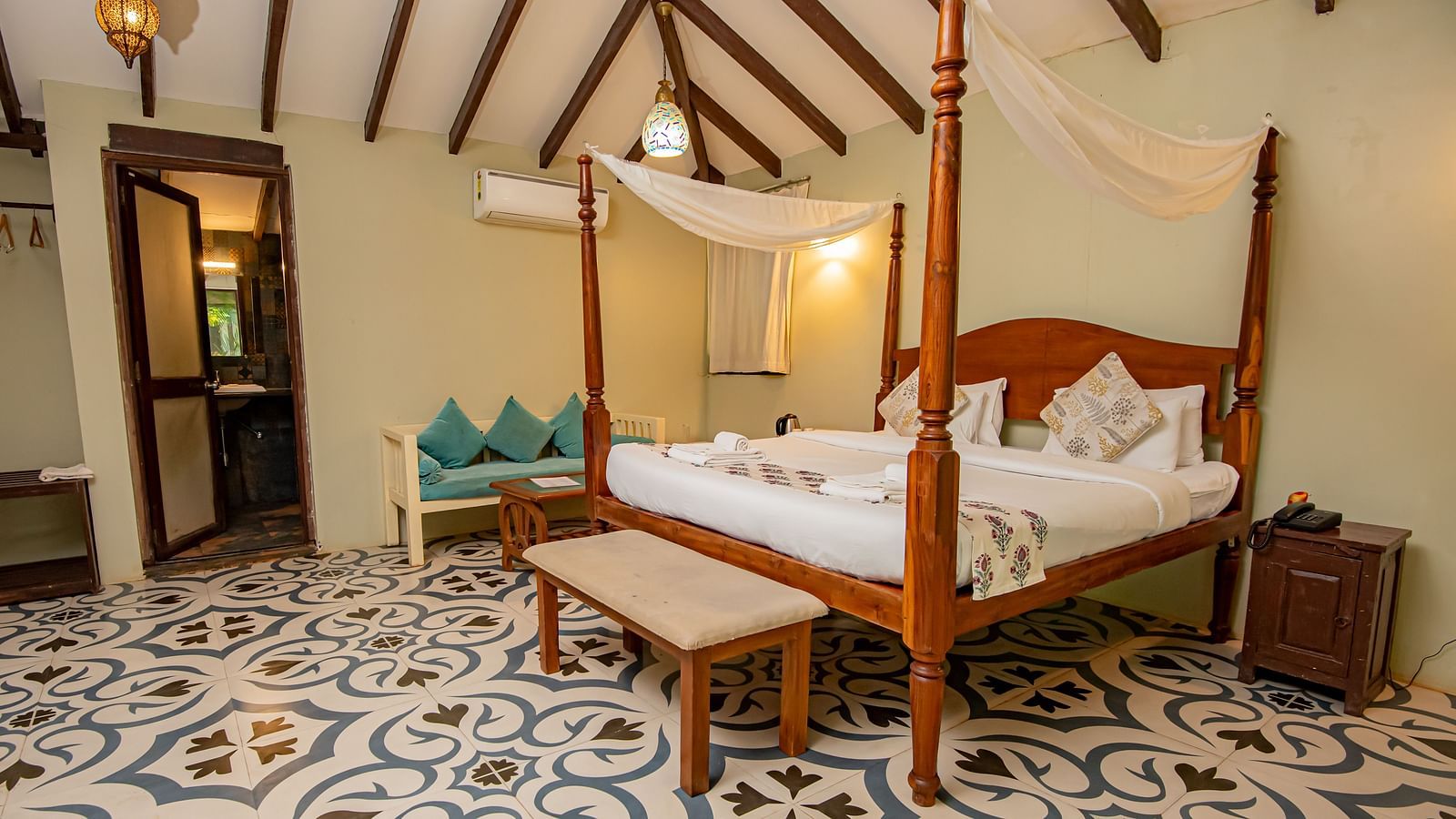 Spacious room with a large bed, wooden seating at the foot, bedside table, and a doorway in the background at De Soul Santé, Morjim.