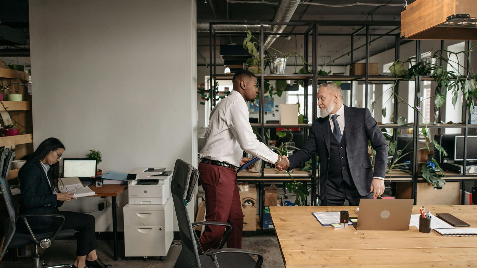 Two men shaking hands while a woman works at a desk in the background in a modern office setting