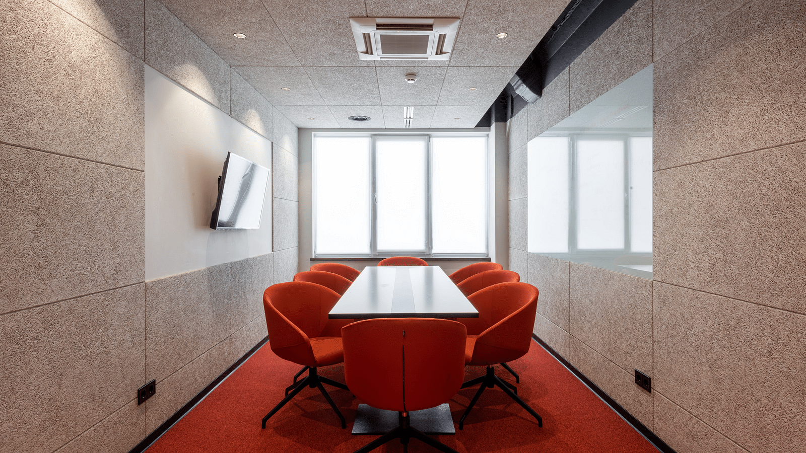 A bright meeting room with large windows, a white and red striped table, surrounded by orange chairs and a TV on a textured wall