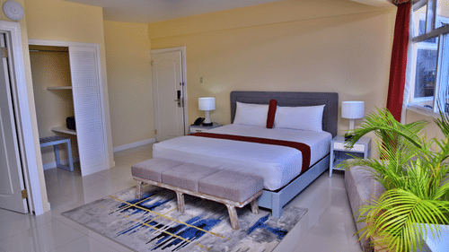 Deluxe Ocean View Room featuring a plush bed along with an ottoman at its foot near the window with plants along the panes and an open wardrobe opposite to it at Hotel Grand A View in Montego Bay