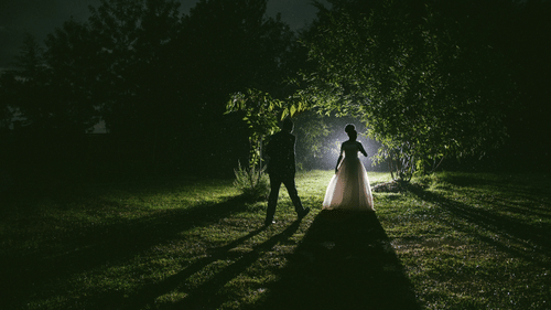 Bride and groom standing in the lawn at night as the light from the background shows the shadow of the bride facing the groom