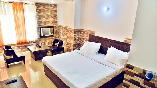 a well decorated room with moderate lighting, a bed with white bedding and chairs at the back in front of a long window covered by curtains from where there is an impression of sunlight  and a television - Clarks Inn Express, Near Jammu Bari Brahmana Railw