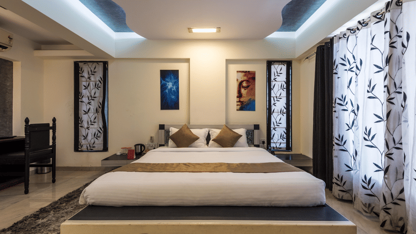 Spacious bedroom with a king size bed, ceiling design lighting, wall art, patterned curtains, and a seating area in the background.