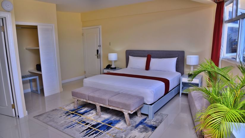 Deluxe Ocean View Room featuring a plush bed along with an ottoman at its foot near the window with plants along the panes and an open wardrobe opposite to it at Hotel Grand A View in Montego Bay