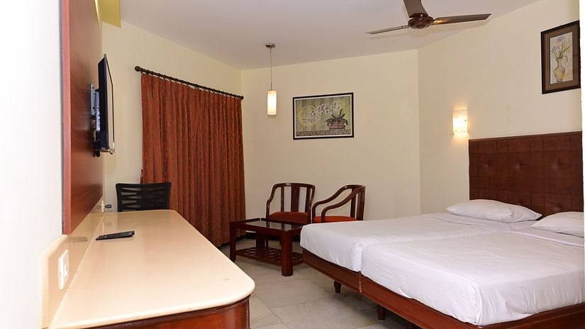 Standard Room with plush bed in front of a TV mounted on the wall and a sitting space by the window featuring two wooden chairs at Jayaram Hotel, Pondicherry
