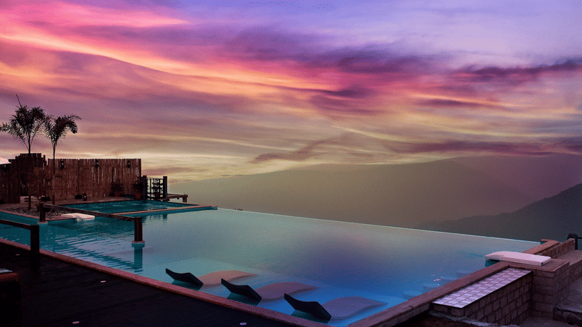 Infinity pool at Polo Resort Cherrapunji at sunset with pink and purple sky, in-water loungers, bamboo screen and palm trees beside mountains.