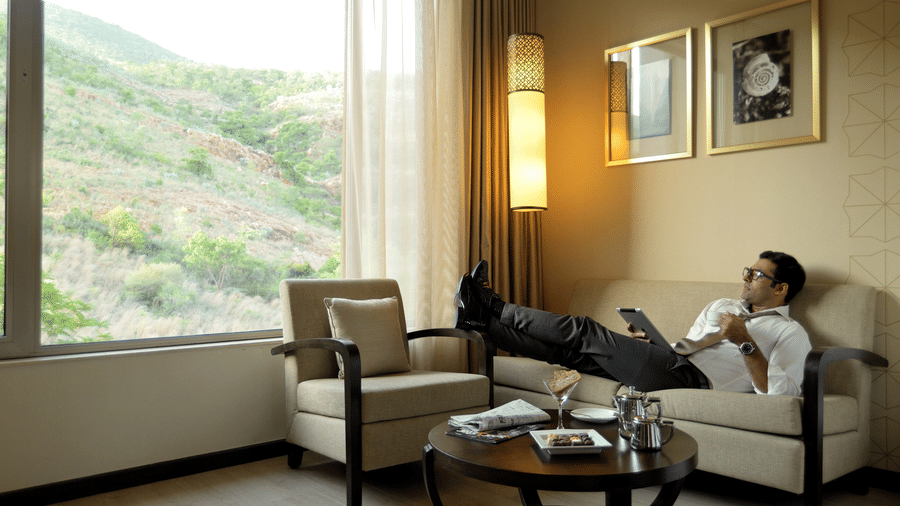 Guest relaxing in the Deluxe Room at Marasa Sarovar Premiere, Tirupati, featuring a comfortable seating area, large picture window with hillside views, and warm contemporary decor.