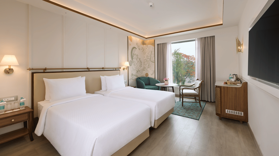 Deluxe Twin Room at Prachi Sarovar Premiere, Bhubaneswar, showing 2 beds, a wall-mounted television, a seating area by the window, and a cabinet.