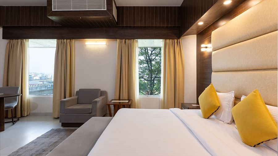 A hotel bedroom featuring a large bed with yellow accent pillows, a dark wood panelled wall behind the headboard, and an armchair by the window - Pride Premier Dehradun