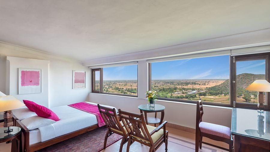 Room with bed, pillows, patterned rug, chairs, table with flowers, desk, lamp, framed artwork, and windows showing outdoor landscape - Tijara Fort-Palace - 19th Century, Alwar.