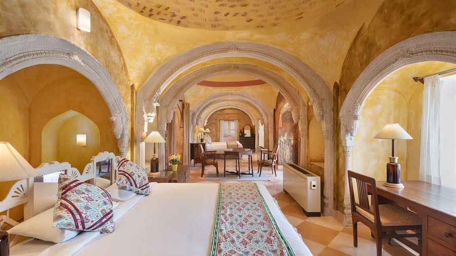Room with bed, patterned pillows, runner, furniture including desk and chairs, arched doorways, patterned ceiling, and visible hallway - Tijara Fort-Palace - 19th Century, Alwar.