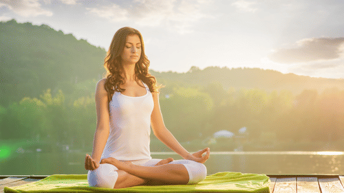 A woman with closed eyes sits in a meditative yoga pose on a green mat on a wooden dock, with a bright sunset over water and hills.