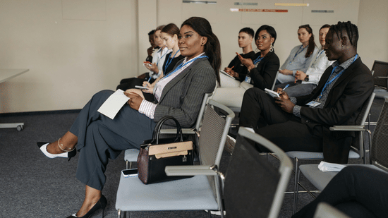 A diverse group of professionally dressed people sitting in rows of chairs, listening to an indoor presentation.