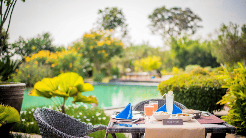 A poolside dining setup surrounded by trees and shrubs at The Serai Bandipur featuring food and beverages kept on the table