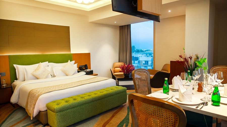 Luxurious hotel room with a king-size bed, modern decor, city view through large window, and an elegant dining setup with bottled water at The Regnant .