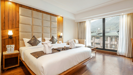 This bright hotel room offers a large bed, wooden floors, and a wide window overlooking a scenic mountain view.