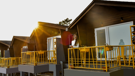 Wooden cottage rooms at Polo Resort Cherrapunji with yellow railing balconies, glass doors and a woman standing on a sunlit terrace.