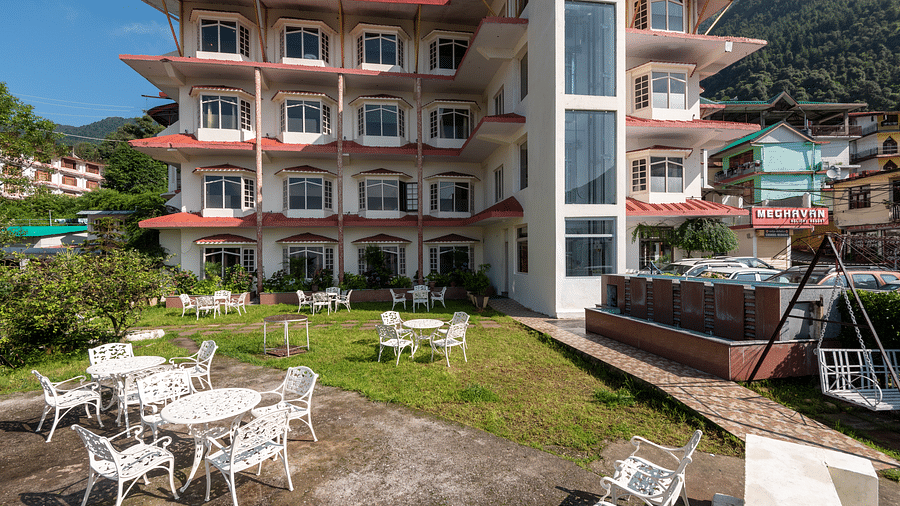 A resort building with a garden, white outdoor tables and chairs, and a swing, set against a hilly backdrop - DLS Meghavan Resort, Dharamshala