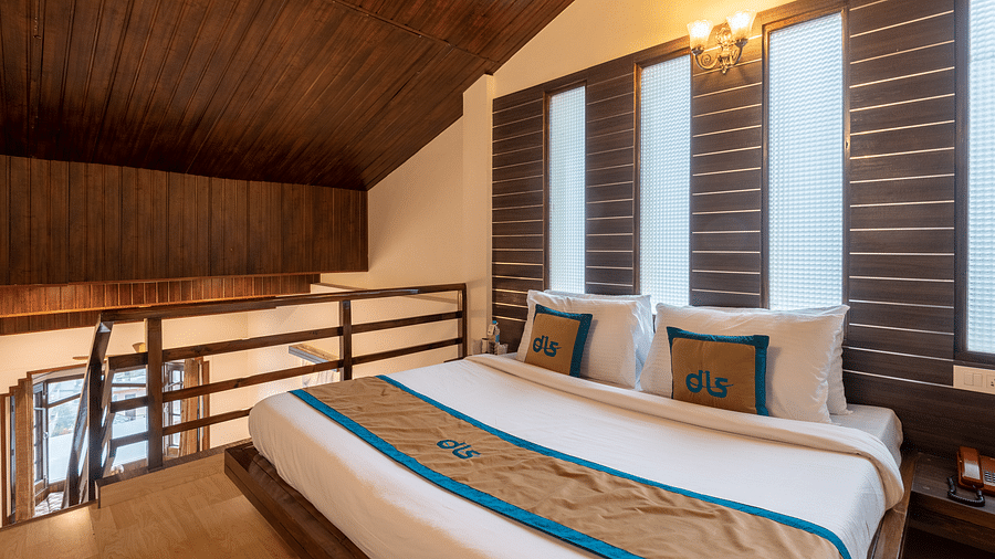 A bedroom with a wooden ceiling, a king size bed with neatly arranged pillows, and a modern design. The room features large windows with blinds and soft lighting - DLS Meghavan Resort, Dharamshala