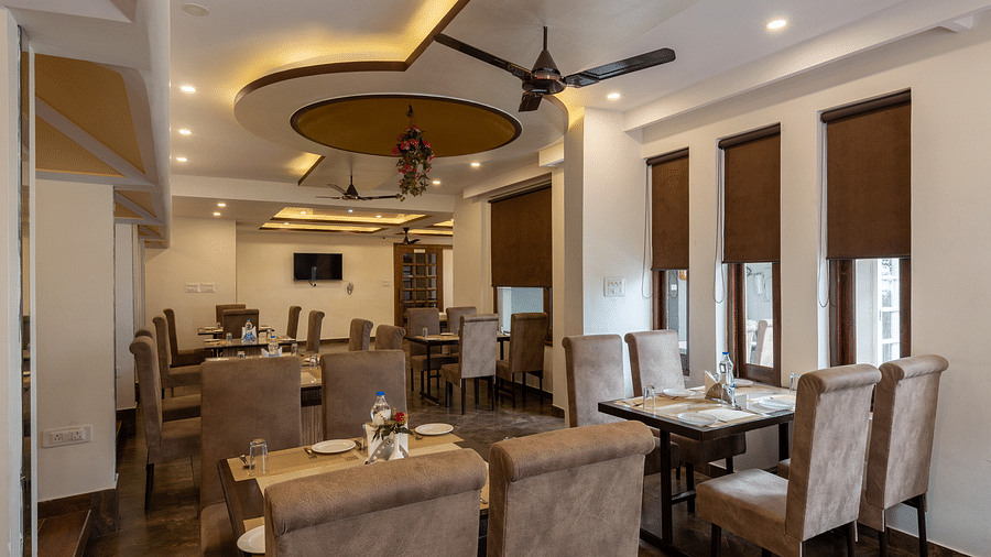 A well-lit dining room with tables and chairs, a ceiling fan, and a TV. The walls are painted white and the ceiling has decorative lighting -  DLS Meghavan Resort