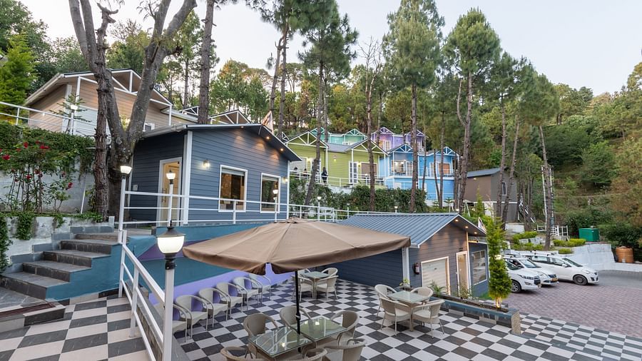 A hillside resort with modern cabins, tall pine trees, and a checkered patio with tables and an umbrella. In the background, terraced buildings and parked white cars are visible - DLS Nature Trinket Resort, Dalhousie.