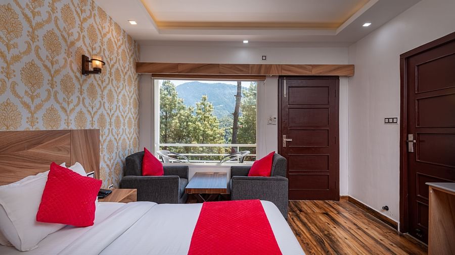  Cosy hotel room with a bed featuring red accents, stylish wallpaper, wooden flooring, seating area by a large window, and views of trees and distant hills - DLS Nature Trinket Resort, Dalhousie.