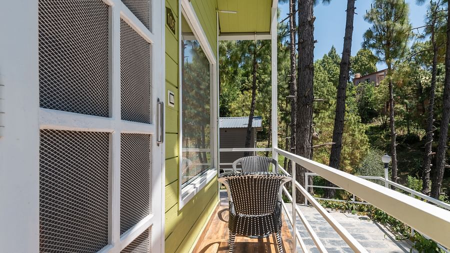  Narrow wooden balcony outside a green cottage with white railings, featuring a small table and chairs, overlooking a peaceful forested area with tall trees - DLS Nature Trinket Resort, Dalhousie.