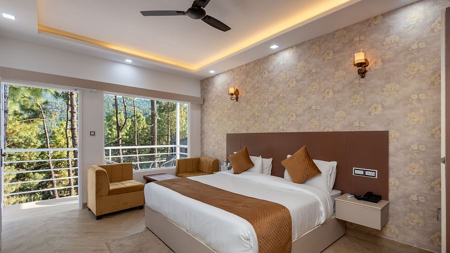 Spacious and well-lit hotel room with a large bed, warm lighting, ceiling fan, and floor-to-ceiling windows offering a view of lush green trees outside - DLS Nature Trinket Resort, Dalhousie.