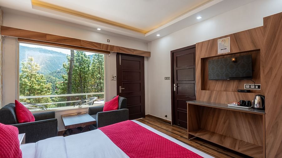 A hotel room with a double bed dressed in white and red linen, a wooden TV unit with a flat-screen, a kettle, and a seating area by a large window offering scenic mountain views - DLS Nature Trinket Resort, Dalhousie.
