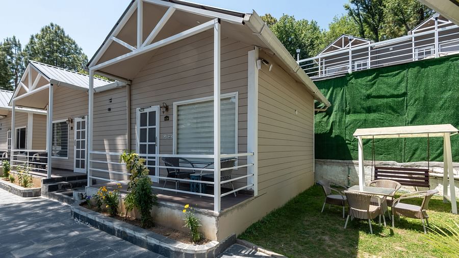  Neatly arranged wooden cottages with front porches along a stone path, featuring outdoor seating and surrounded by trees and greenery under a sunny sky - DLS Nature Trinket Resort, Dalhousie.