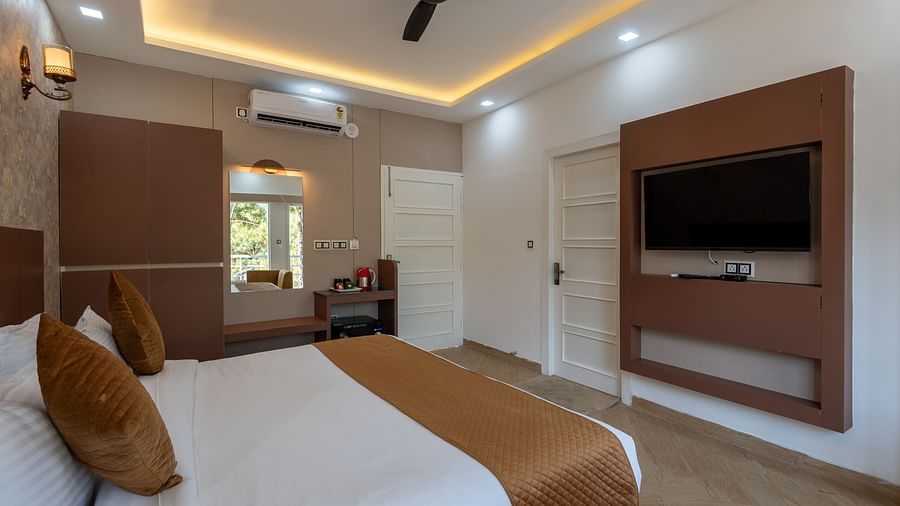 Modern hotel room with a double bed, brown and white bedding, wall-mounted TV, ceiling fan, and a dressing table near a window with natural light - DLS Nature Trinket Resort, Dalhousie.