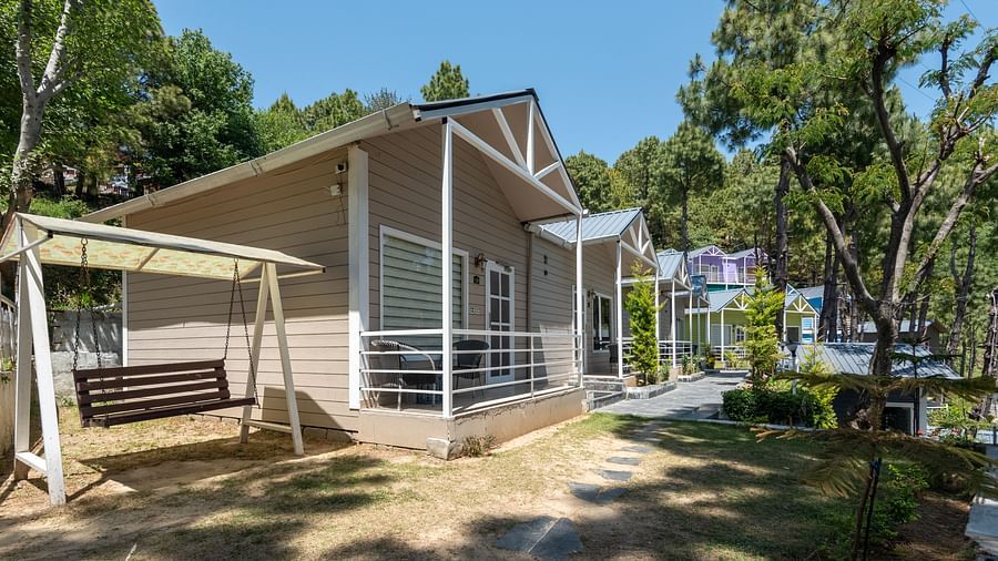A charming cottage with a sloped roof and large windows, featuring a swing on the porch and a pathway leading through a lush green garden, surrounded by tall trees - DLS Nature Trinket Resort, Dalhousie.