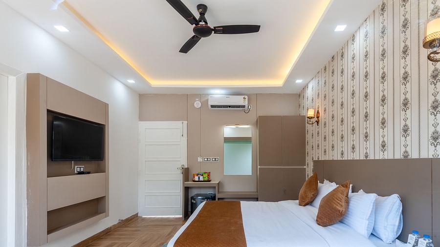 Stylish hotel room featuring a large bed with decorative pillows, wall-mounted TV, ceiling fan, ambient lighting, and a vanity area with a mirror and minibar - DLS Nature Trinket Resort, Dalhousie.