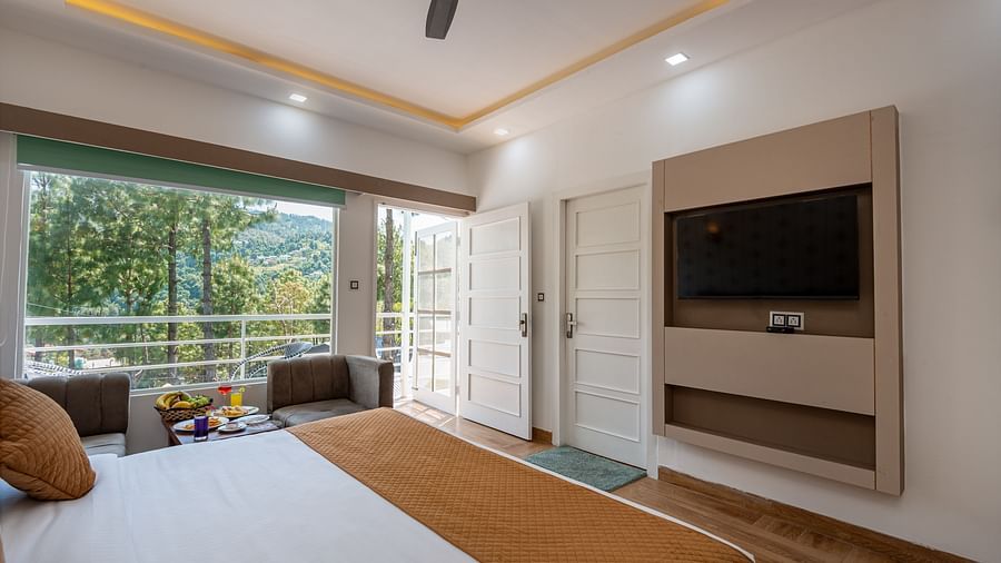 A bright hotel room with a double bed, TV, and seating area, opening onto a balcony with scenic hill views - DLS Nature Trinket Resort, Dalhousie.