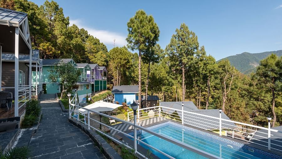 Daytime view of a serene hillside resort with a swimming pool, colourful cabins, pine trees, and mountain scenery in the background, alongside a stone walkway - DLS Nature Trinket Resort, Dalhousie.