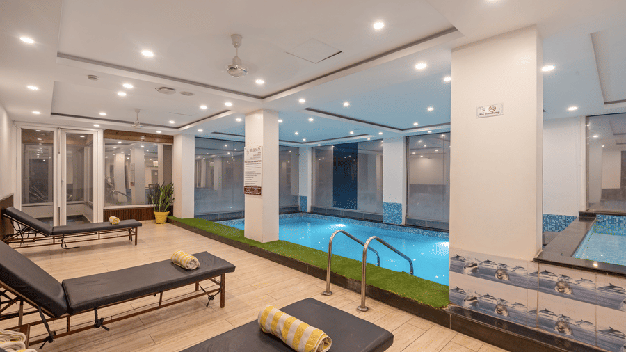 A dining area next to the indoor swimming pool at DLS Hotels On The Ganges. A square wooden table with four chairs is set with plates and glasses of juice. The pool is visible in the background, with its blue mosaic tiling. A modern chandelier hangs from